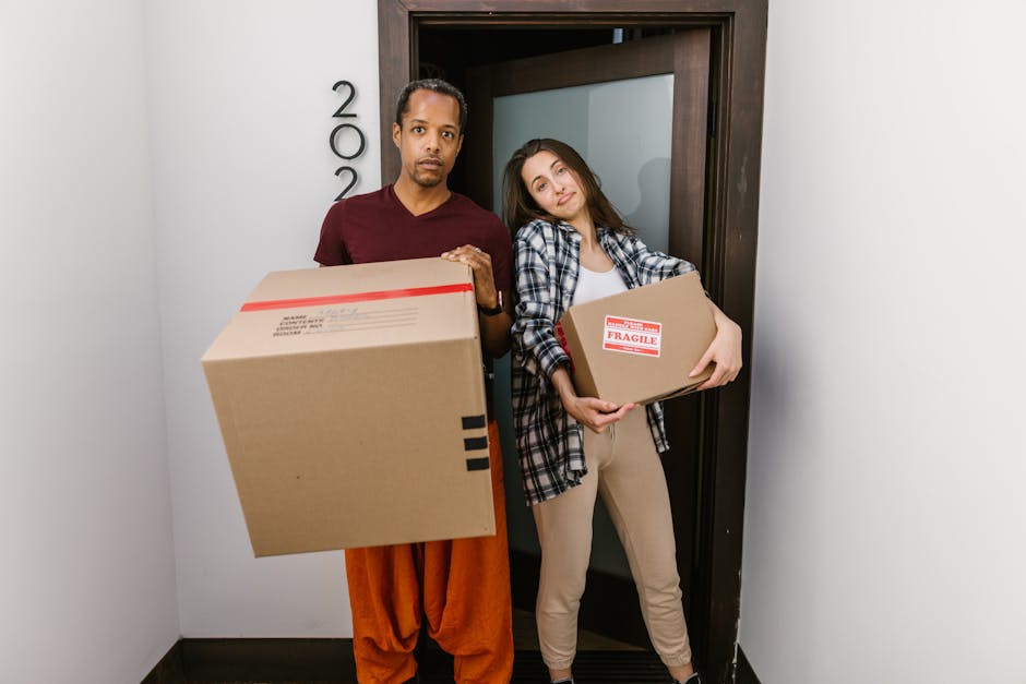 A man and a woman stand inside a building near an entrance door, each holding a cardboard box used for packing household items during home relocation. The man, dressed in a maroon t-shirt and orange t