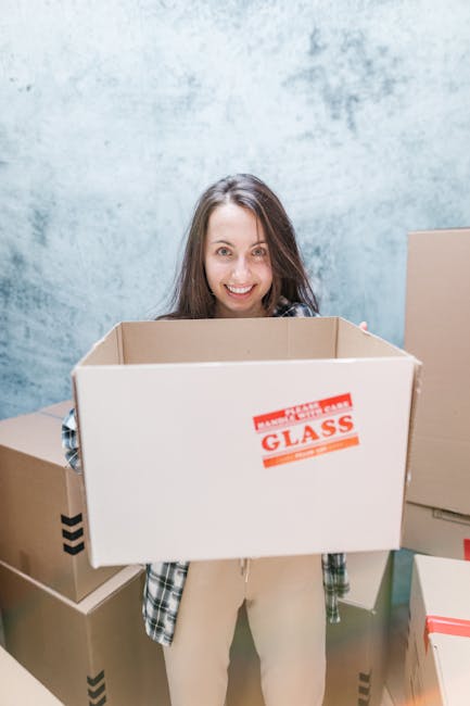 A man and a woman stand inside a building near an entrance door, each holding a cardboard box used for packing household items during home relocation. The man, dressed in a maroon t-shirt and orange t