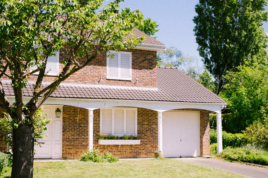 A residential street scene showing a row of modern brick houses with pitched roofs, some with dormer windows, situated along a curved road with well-maintained lawns and small trees in front gardens. 