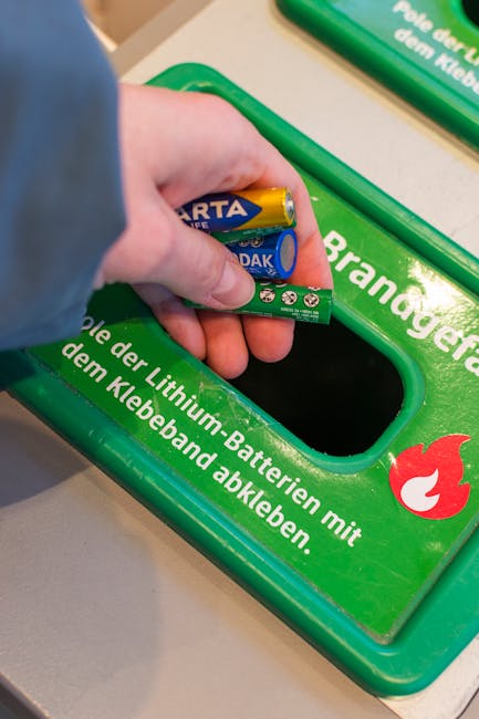 A person wearing a white shirt and gloves is holding a small cardboard box above a blue recycling bin with a white recycling symbol on the front, set against a light green wall. The recycling bin is p