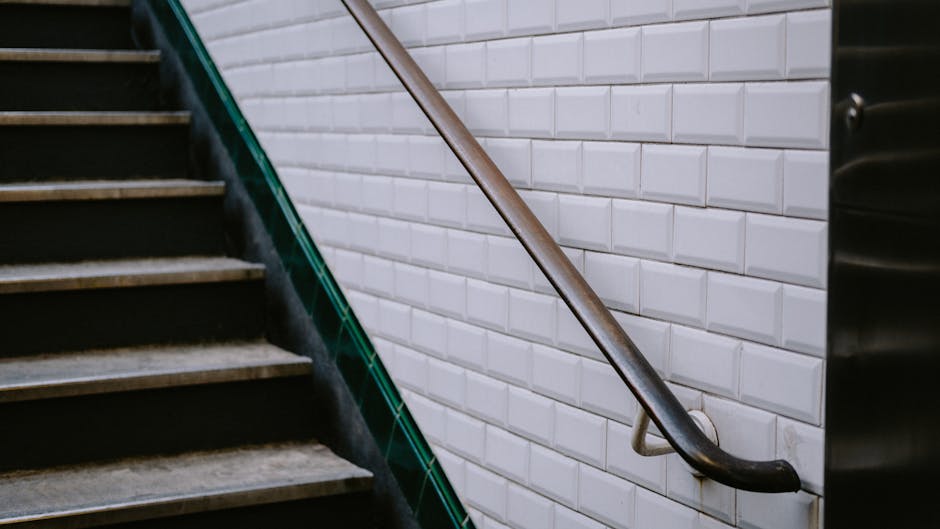 A concrete and metal staircase with wooden steps and blue railings leading from a sidewalk to an elevated walkway between modern glass and brick office buildings in an urban setting. The staircase is