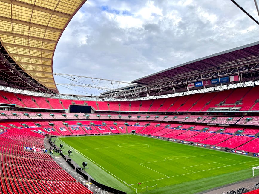 Interior view of a large sports stadium with bright red seating arranged in multiple tiers around a well-maintained green football pitch. The stadium is partially covered by a curved roof structure wi