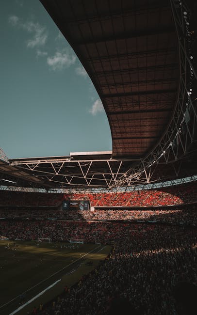 Interior view of a large sports stadium with bright red seating arranged in multiple tiers around a well-maintained green football pitch. The stadium is partially covered by a curved roof structure wi