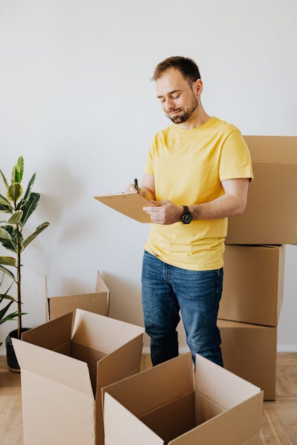 A man and a woman are seen carrying cardboard boxes during a house relocation process outside a modern residential property. The woman, standing inside the doorway, holds a medium-sized cardboard box 